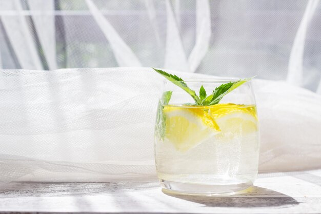 Primer plano de limonada casera con menta de limón y cubitos de hielo en un vaso sobre una mesa de madera blanca con tela de tul blanco