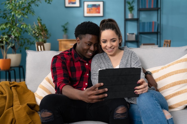 Primer plano de una linda y alegre pareja casada multiétnica descansando en un sofá en una acogedora habitación hablando por videochat