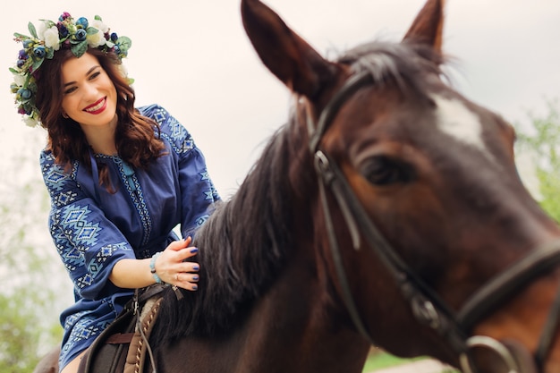 Primer plano de una linda chica con una corona de flores en la cabeza y un hermoso maquillaje que se sienta en un caballo y sonríe francamente