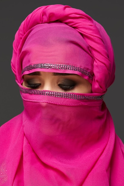 Primer plano de una linda joven con expresivos ojos ahumados que lleva un elegante hiyab rosa decorado con lentejuelas. Ha vuelto la cabeza y ha cerrado los ojos un fondo oscuro. Emociones humanas,