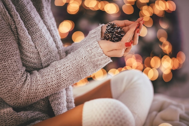 Primer plano de una linda joven sosteniendo piñas en sus manos rodeada de luces navideñas.