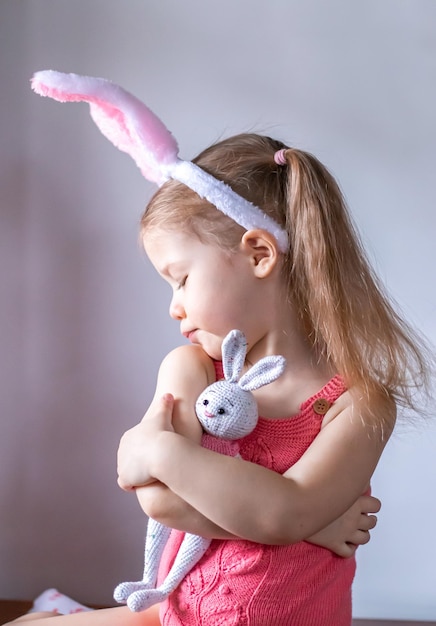 Primer plano de una linda niña con orejas de conejo de Pascua abrazando un conejito de juguete sobre un fondo claro