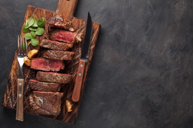 Primer plano listo para comer bistec Nueva York razas de carne de Angus negro con hierbas ajo y mantequilla en un tablero de madera El plato terminado para la cena en un fondo de piedra oscura Vista superior con espacio de copia
