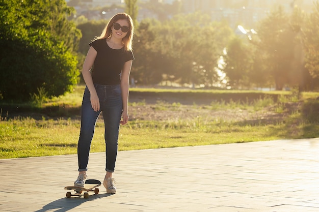 Primer plano de longboard monopatín en manos de una mujer joven en jeans y una camiseta negra en el parque.