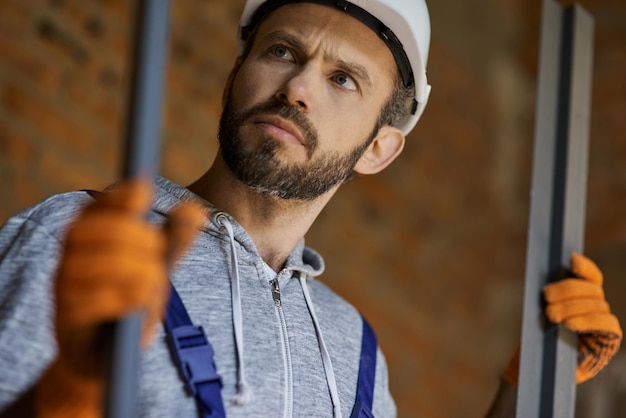 Foto primer plano de macho joven constructor con casco mirando enfocado sosteniendo espárragos metálicos