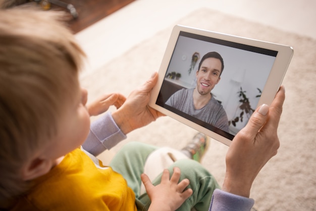 Primer plano de la madre sosteniendo la tableta y mostrando al hijo mientras conecta al padre a través de la aplicación de videoconferencia