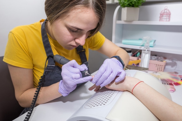 Primer plano de una maestra haciendo una manicura a un cliente usando una máquina de manicura. Una manicurista trabaja con un taladro eléctrico para uñas en un salón de belleza. Proceso de hardware de manicura.