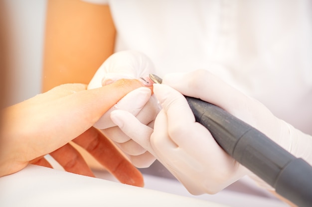 Primer plano de maestro de manicura en guantes blancos aplicando un taladro de lima de uñas eléctrica para quitar el esmalte de uñas en el salón de belleza.