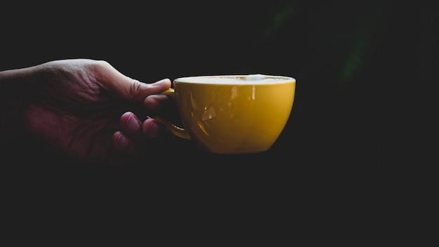 Primer plano mano aislada sosteniendo la taza amarilla sobre fondo negro. Asimiento de la mano simple taza amarilla. Perfecto tanto para té como para café.
