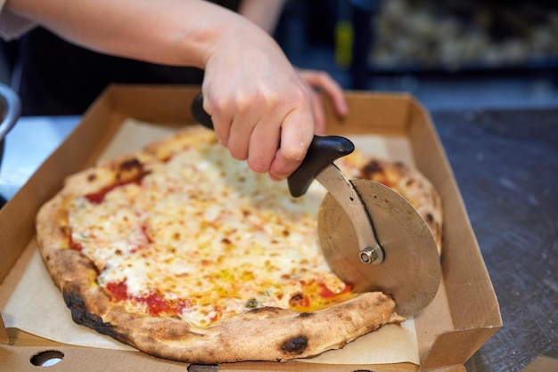 Foto primer plano mano de chef panadero en uniforme blanco cortando pizza en la cocina.