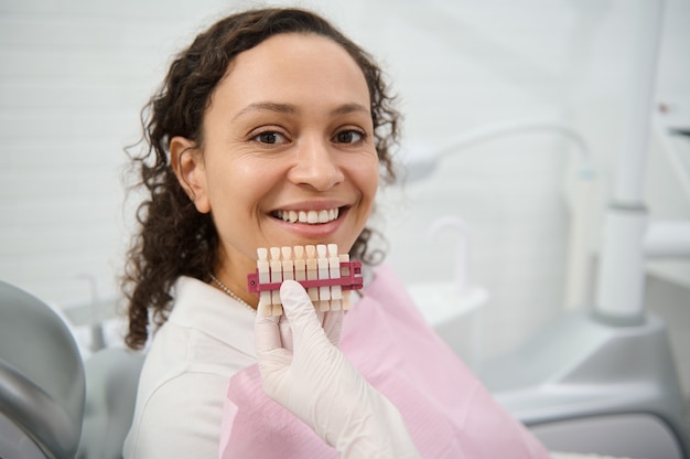 Primer plano de la mano del dentista sosteniendo una carta de colores de los dientes cerca de la cara de una clínica de odontología paciente mujer bonita sonriendo con hermosa sonrisa con dientes mirando a la cámara. Blanqueamiento de dientes, concepto de blanqueamiento