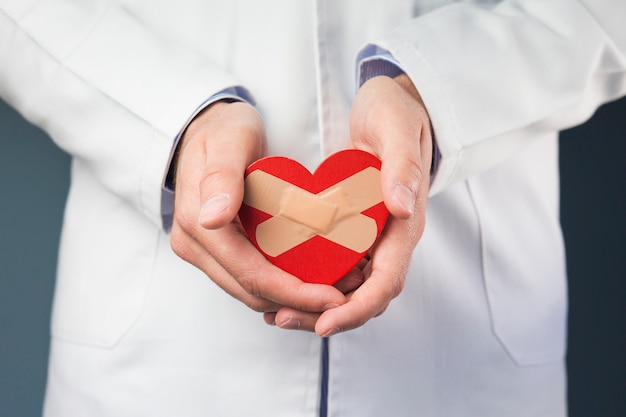 Foto primer plano de la mano del doctor sosteniendo corazón rojo con vendas cruzadas