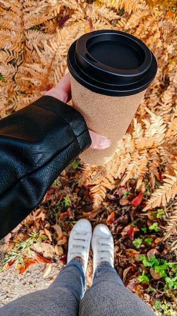 Primer plano de una mano femenina joven sosteniendo una taza de café contra el fondo del follaje de otoño