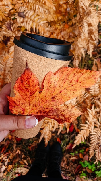 Primer plano de una mano femenina joven sosteniendo una taza de café contra el fondo del follaje de otoño