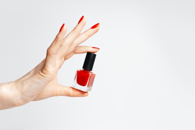 Primer plano de una mano femenina con manicura roja sosteniendo un esmalte de uñas brillante de color rojo sobre fondo blanco