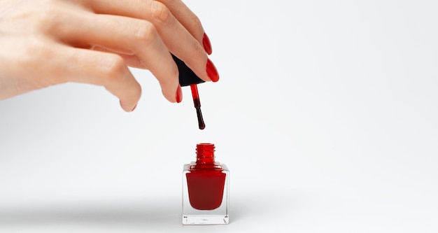 Primer plano de una mano femenina con manicura roja sosteniendo un esmalte de uñas brillante de color rojo sobre fondo blanco