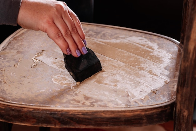 Primer plano de la mano femenina que pone masilla y nivela la superficie de la silla envejecida preparando un mueble para colorear más en el taller doméstico para la renovación