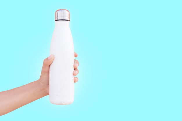 Primer plano de la mano femenina que sostiene la botella de agua termo de acero reutilizable blanca aislada en la superficie de color cian con espacio de copia.
