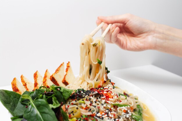 Primer plano de mano femenina sosteniendo palillos con fideos en sopa de ramen asiático