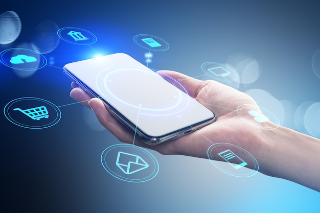 Primer plano de una mano femenina sosteniendo un teléfono inteligente blanco con una interfaz digital redonda creativa con varios iconos en un fondo azul borroso Transformación digital concepto de compras y almacenamiento bancario en línea