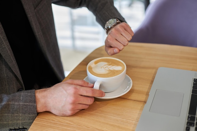 Primer plano de la mano de un hombre de negocios sosteniendo una taza de café, revisando el tiempo mirando el reloj sentado en el café