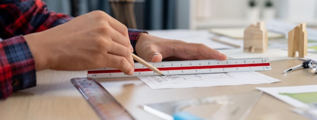 Foto primer plano de la mano del ingeniero arquitecto usando una regla para madurar y dibujar un plano en la mesa de reuniones con lápiz de bloque de madera y planos dispersos en la oficina moderna de arquitectura delineación