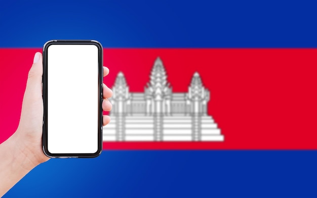 Primer plano de la mano masculina que sostiene el teléfono inteligente con pantalla en blanco en el fondo de la bandera borrosa de Camboya