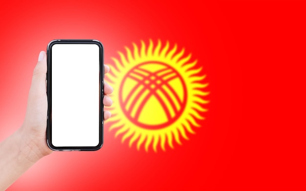 Primer plano de la mano masculina que sostiene el teléfono inteligente con pantalla en blanco en el fondo de la bandera borrosa de Kirguistán