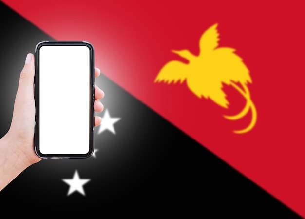 Primer plano de la mano masculina que sostiene el teléfono inteligente con pantalla en blanco en el fondo de la bandera borrosa de Papúa Nueva Guinea