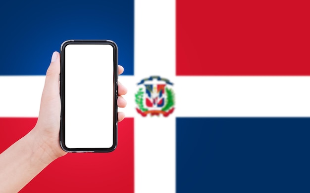 Primer plano de la mano masculina que sostiene el teléfono inteligente con pantalla en blanco en el fondo de la bandera borrosa de República Dominicana