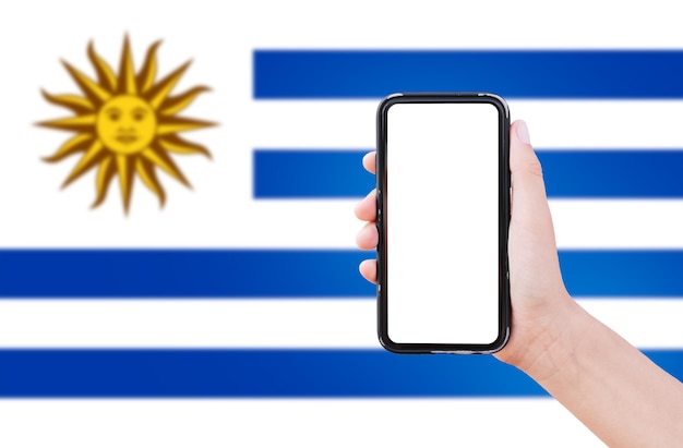 Primer plano de la mano masculina que sostiene el teléfono inteligente con pantalla en blanco en el fondo de la bandera borrosa de Uruguay