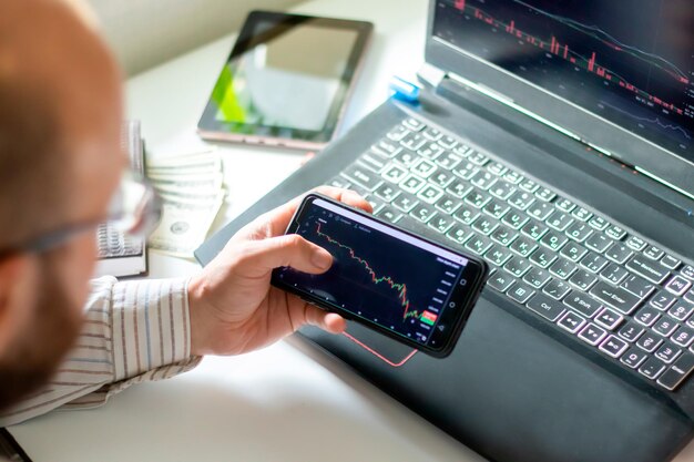 Primer plano de la mano masculina que sostiene el teléfono móvil con el stock de monitoreo de gráfico financiero. Comercio en línea