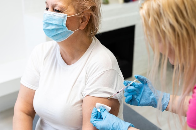 Primer plano de la mano del médico general que sostiene la inyección de la vacuna mientras usa una máscara protectora facial durante la pandemia de covid-19. Enfermera joven con mascarilla quirúrgica inyectando a una anciana.