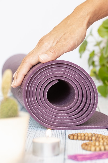 Primer plano de la mano de una mujer desplegando una estera de yoga violeta, cuentas de madera en madera blanca. Accesorios esenciales para practicar yoga y meditación.