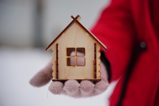 Primer plano de la mano de la mujer en el guante sosteniendo una pequeña casa de madera en invierno Concepto de compra de un nuevo apartamento