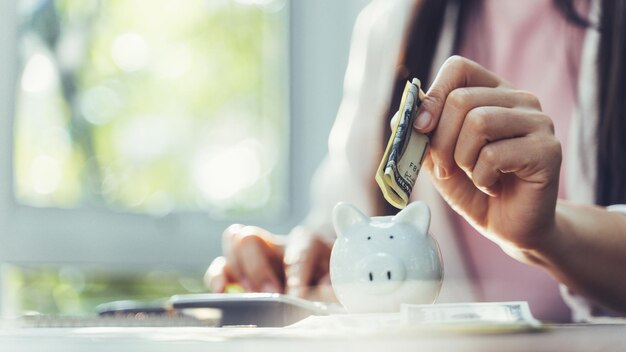 Primer plano de la mano de una mujer de negocios poniendo billetes de dinero en una alcancía para ahorrar dinero