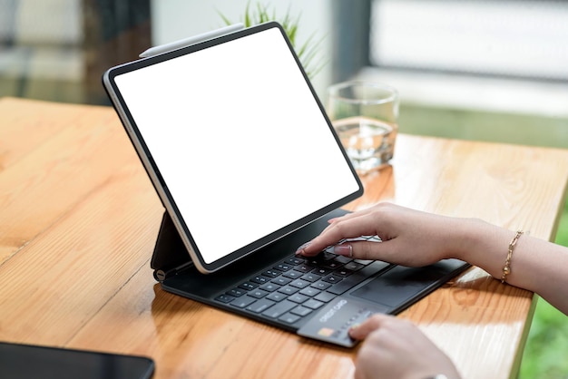 Primer plano de la mano de la mujer usando la pantalla blanca en blanco de la tableta de la computadora y sosteniendo compras con tarjeta de crédito en línea o banca por Internet en el escritorio de la oficina.