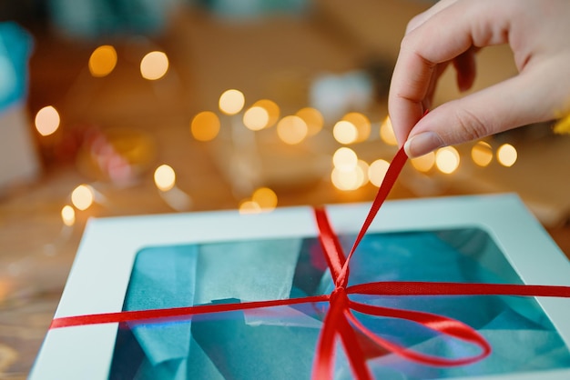 Primer plano de la mano de la niña tirando de la cinta roja de las luces bokeh de regalo en el fondo desembalaje de la caja en woode ...