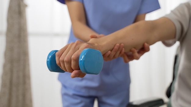 Foto primer plano de la mano de una persona discapacitada que pasa por un entrenamiento de rehabilitación usando una mancuerna. ayudante de enfermería femenina recortada proporcionó asistencia en el fondo