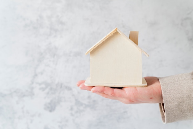 Primer plano de la mano de la persona de negocios que sostiene el modelo de casa de madera en miniatura contra el muro de hormigón blanco