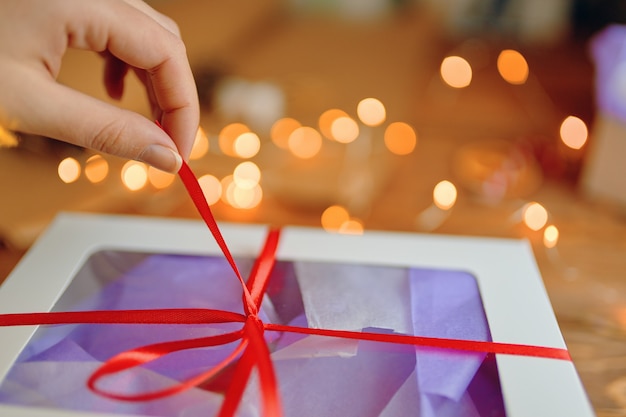 Primer plano de la mano de las personas tirando de la cinta roja del concepto festivo de regalo de navidad o cumpleaños bokeh ...