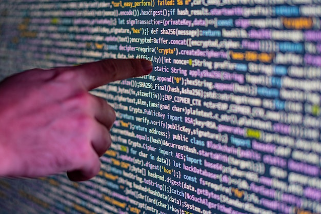 Primer plano de una mano que señala sobre una gran pantalla de visualización brillante llena de código colorido que posiblemente representa la programación de computadora o la visualización de datos