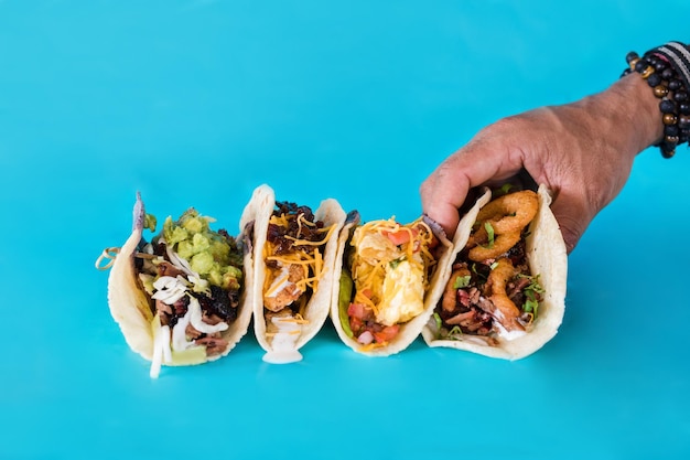 Primer plano de una mano recogiendo tacos gourmet con diferentes rellenos sobre una superficie azul
