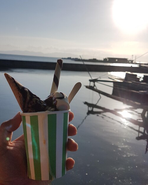 Foto primer plano de la mano sosteniendo el helado contra el mar durante la puesta de sol