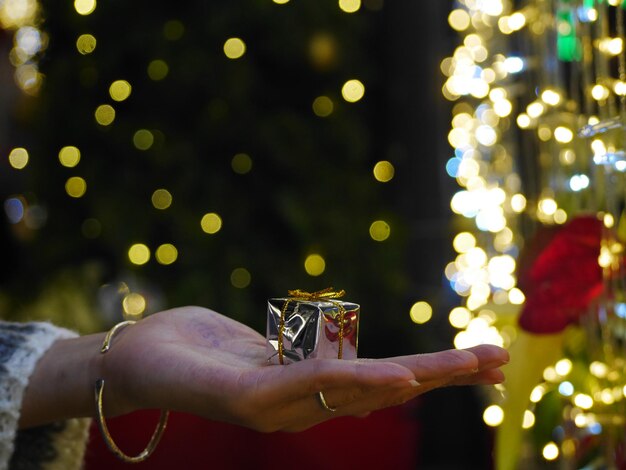 Foto primer plano de la mano sosteniendo un regalo contra el árbol de navidad iluminado por la noche