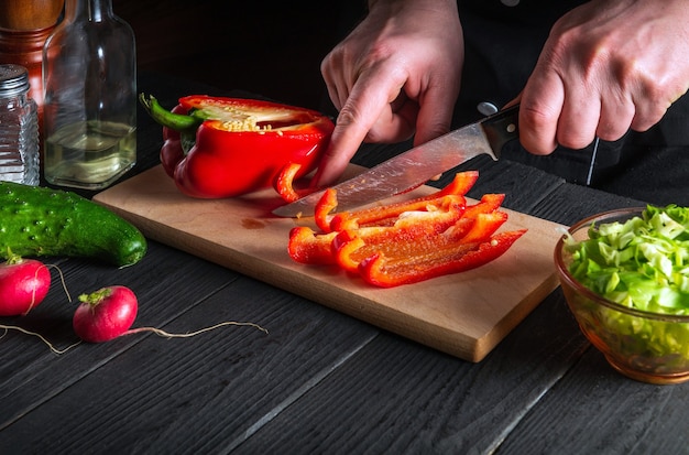 Primer plano de las manos del cocinero o del cocinero cortando pimientos en la tabla de cortar. Preparación profesional de ensalada en la cocina de un restaurante o cafetería.