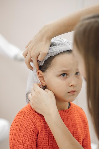Primer plano de las manos de esteticista poniendo una gorra médica desechable blanca en la cabeza de una niña de seis años en la oficina de cosmetología preparándose para la perforación del oído