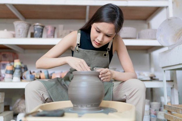 Primer plano de las manos femeninas del artista asiático ceramista trabajando en alfareros mujer escultura femenina asiática rueda con manos sucias mojadas formando jarrón de arcilla en la rueda de cerámica en el estudio del taller