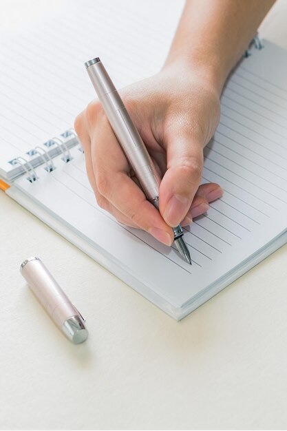 Un primer plano de manos femeninas escribiendo en un bloc de notas en espiral colocado en un escritorio blanco usando un bolígrafo de tinta