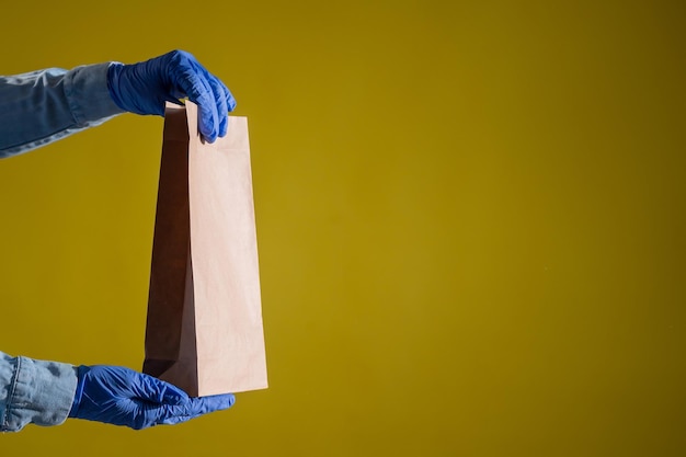 Primer plano de manos femeninas con guantes y una camisa de mezclilla El repartidor sostiene una bolsa de papel vacía sobre un fondo amarillo Embalaje artesanal para bocadillos para llevar